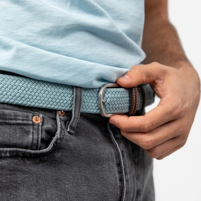 Person adjusting a blue woven belt with a silver buckle on gray jeans against a white background